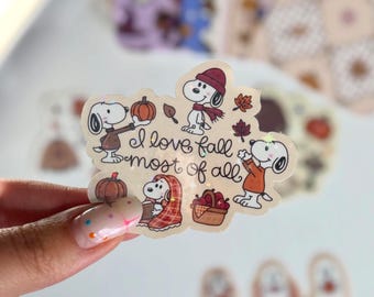 May include: A die-cut sticker featuring Snoopy in various autumn-themed poses, including holding a pumpkin, wearing a scarf, and wrapped in a plaid blanket. The text "I love fall most of all" is written in black script in the centre with small stars.