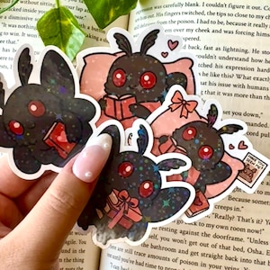 Può includere: Quattro adesivi glitter neri e rossi con una falena dei cartoni animati che tiene un libro, un libro con un cuore, una pila di libri con un fiocco e un libro con il testo "Have You Seen Me?" Gli adesivi sono su un libro rosa e bianco.