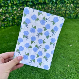 May include: A rectangular card with a floral pattern featuring blue hydrangea blooms, green leaves, and small bows on a white background. The card is held in a person's hand, with a green lawn and foliage in the background.