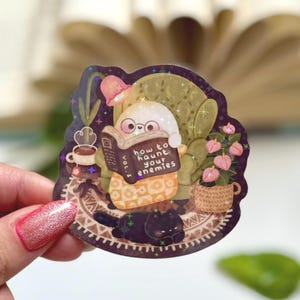 May include: A die-cut sticker featuring a small white ghost with glasses sitting in a green armchair, reading a book titled How to Haunt Your Enemies Vol. I. A black cat lies on a patterned rug nearby, alongside a potted plant and a cup of tea.