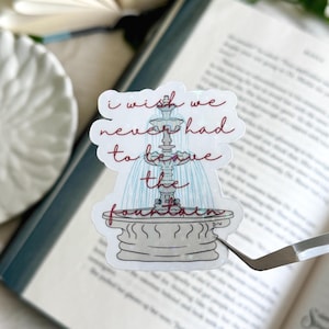 May include: A sticker featuring a fountain illustration with the text "I wish we never had to leave the fountain" in red script. The sticker is on a book page, with a white daisy-shaped plate in the background.