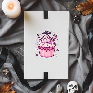 May include: A white card featuring a pink cupcake illustration. The cupcake has pink frosting, sprinkles, and a bat on top. The card rests on grey fabric with a black ribbon, a candle, and decorative items.