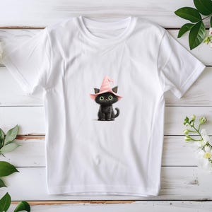 May include: White t-shirt with a black cat illustration wearing a pink witch's hat. The cat has large green eyes. The design is a whimsical illustration, suitable for Halloween or cat enthusiasts.