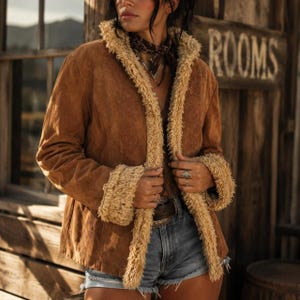 May include: A tan suede jacket featuring a textured beige faux-shearling trim along the front opening and sleeve cuffs. The jacket is worn over blue denim shorts and includes a dark brown leather belt with a metal buckle visible at the waist.