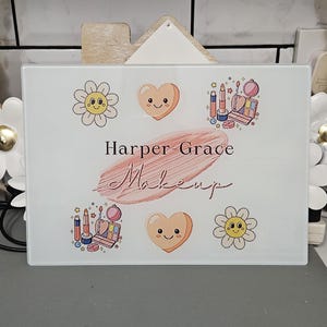 May include: White rectangular sign featuring the text "Harper Grace Makeup". The sign is decorated with cartoon hearts, flowers, and makeup illustrations in pink, orange, and blue.
