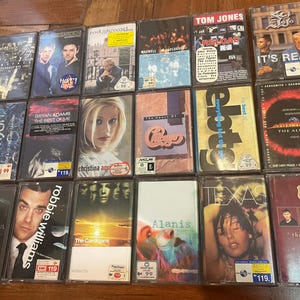 May include: A collection of 12 cassette tapes with various artists and album titles. The tapes are arranged in three rows of four. The tapes are all black with white text and images. Some of the artists include Tom Jones, Bryan Adams, Robbie Williams, and Alanis Morissette.