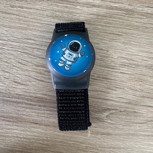 Könnte beinhalten: Ein schwarzes Armband mit einem kreisförmigen, blauen Zifferblatt mit einem Cartoon-Astronauten in einem weißen Raumanzug. Das Armband hat ein schwarzes, gewebtes Armband und ein dunkelgraues Kunststoffgehäuse.