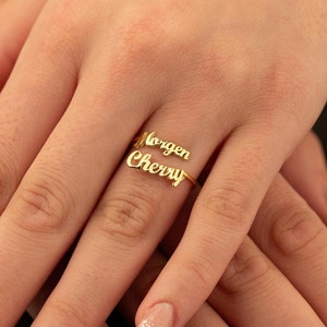 May include: Gold ring with the names "Morgan" and "Cherry" written in cursive on it. The ring is worn on the ring finger of a hand with pink glitter nail polish.