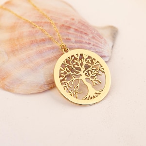 May include: A gold-colored necklace featuring a circular pendant with a detailed tree of life design. The pendant hangs from a delicate chain. The necklace is displayed on a seashell against a white background.
