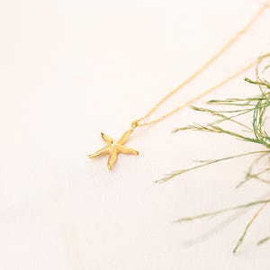 May include: A gold chain necklace with a delicate starfish pendant. The necklace is photographed against a white background with green foliage.