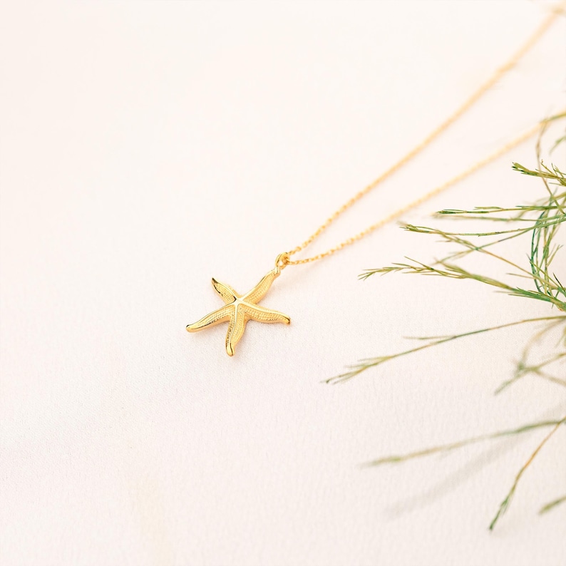May include: A gold chain necklace with a delicate starfish pendant. The necklace is photographed against a white background with green foliage.