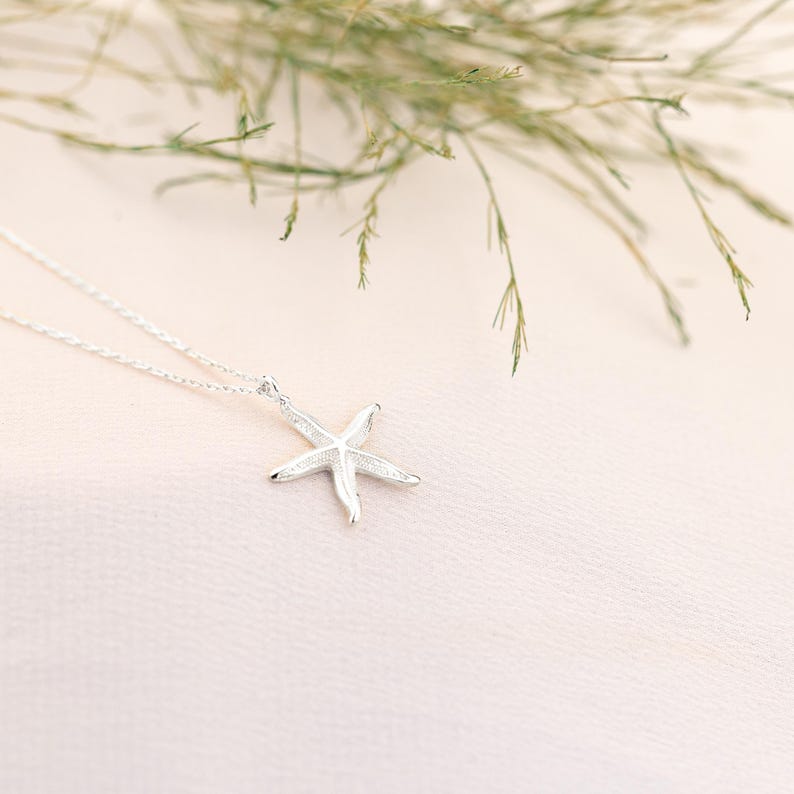 May include: A silver necklace with a delicate starfish pendant. The necklace is on a pink background with green foliage.