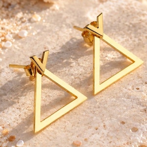 Peut inclure: Une paire de boucles d'oreilles à tige dorées présentant un design en forme de triangle ouvert. Chaque boucle possède un petit accent en V sur le dessus où la tige est fixée, créant une silhouette géométrique avec une finition métallique polie.