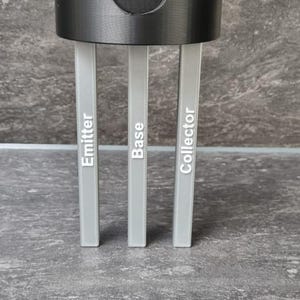 Könnte beinhalten: Eine schwarze, zylindrische Komponente mit einer kreisförmigen Öffnung, verbunden mit drei grauen, rechteckigen Stiften. Die Stifte sind mit "Emitter", "Base" und "Collector" beschriftet. Das Objekt befindet sich auf einer grauen, strukturierten Oberfläche.