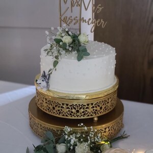 Könnte beinhalten: Eine zweistöckige Hochzeitstorte mit weißem Frosting und einer Wolfsfigur auf der unteren Etage. Die Torte ist mit kleinen weißen Punkten verziert und mit einem Holzschild mit der Aufschrift "Mr. & Mrs. Kassmeyer" gekrönt.