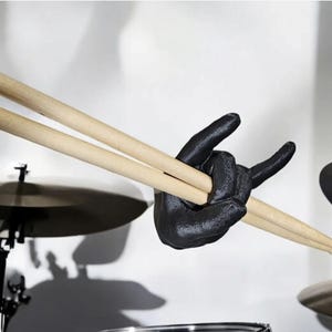 May include: A black hand-shaped drumstick holder in the "rock on" gesture, holding two wooden drumsticks. The background includes drum cymbals and stands, suggesting a music-themed product.