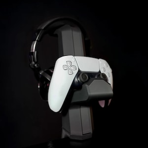 May include: A black and gray gaming headset and a white and gray game controller are displayed on a black stand. The controller is in the foreground, with the headset behind it. The stand is black.