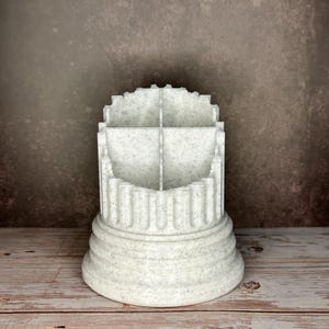 May include: A white speckled desk organizer shaped like a classical column with a tiered, circular base and a fluted exterior. The top section is divided into four distinct compartments by intersecting walls, creating a functional storage container.