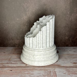 May include: A white speckled desk organizer shaped like a broken classical column. The object features a wide, tiered circular base and vertical fluted detailing along the exterior. The top edge has a jagged, uneven finish resembling stone.
