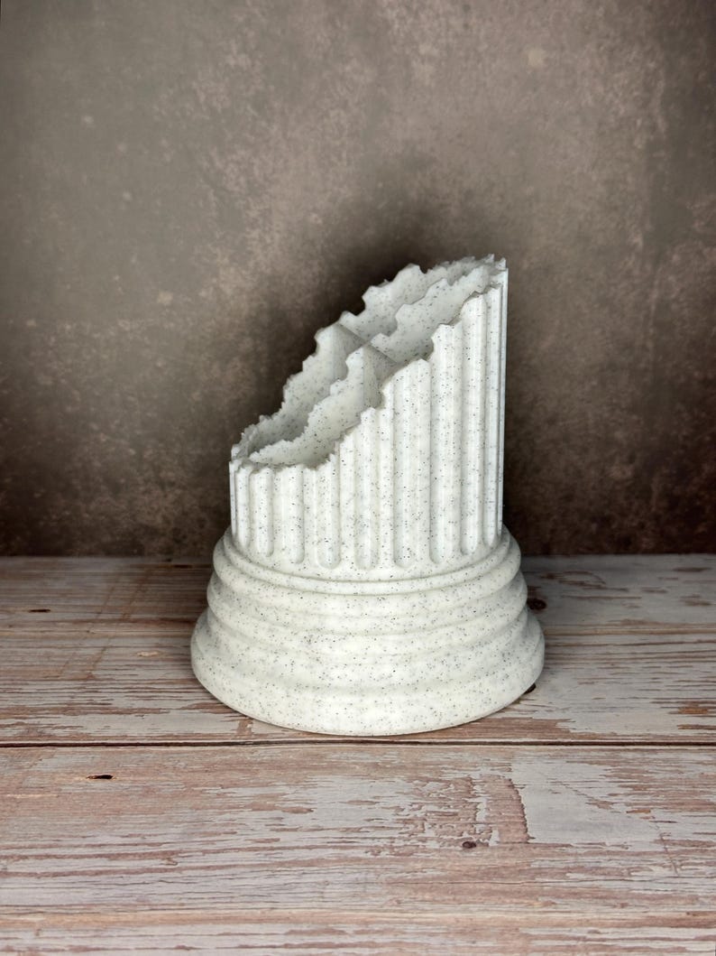May include: A white speckled desk organizer shaped like a broken classical column. The object features a wide, tiered circular base and vertical fluted detailing along the exterior. The top edge has a jagged, uneven finish resembling stone.