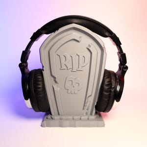 May include: A gray tombstone-shaped headphone stand with the words "RIP" and a skull design. Black headphones are resting on the stand.