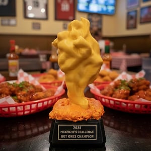 May include: A unique trophy with a yellow, abstract shape on top of a base. The base is black with a gold plaque that reads "2025 McKenzie's Challenge Hot Ones Champion". Red baskets of food are in the background.