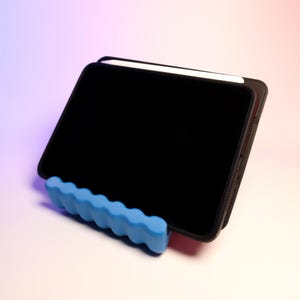 May include: A black tablet computer with a white stylus resting on top of it. The tablet is propped up on a blue, wavy stand.