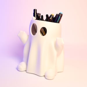 May include: White ghost-shaped ceramic pencil holder with a variety of pens and pencils sticking out of the top.