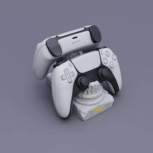 May include: Two white and black wireless game controllers featuring the Sony logo on the top edge. The controllers are positioned on a small, white, stone-like pedestal base that has a single yellow lightning bolt icon on the front side.