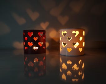 May include: Two cylindrical candle holders, one dark red and one white, each featuring a repeating pattern of heart-shaped cutouts. Light shines through the cutouts, casting heart-shaped shadows on the wall and reflecting on the surface.