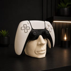 May include: A white and black video game controller resting on a beige human face sculpture. The controller features a directional pad, two analog sticks, and action buttons. The sculpture depicts a nose, mouth, and chin, serving as a stand.