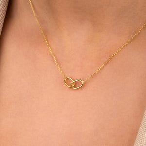 Op de afbeelding: Een delicate gouden ketting met twee in elkaar grijpende cirkels. De hanger hangt aan een fijne ketting, waardoor een minimalistisch en elegant ontwerp ontstaat. De ketting wordt gepresenteerd tegen een neutrale achtergrond.