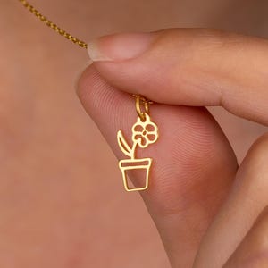 May include: A delicate gold-colored necklace featuring a pendant shaped like a potted flower. The pendant is a simple outline of a flower in a pot. The necklace chain is also gold-colored and appears to be a delicate link chain.