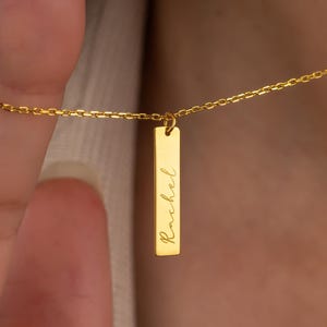 Puede incluir: Un collar dorado con una cadena fina y un colgante rectangular. El colgante lleva grabado el nombre "Rachel" en escritura cursiva. El collar se muestra sobre un fondo neutro.