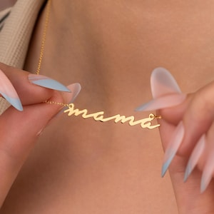 以下が含まれることがあります： 筆記体で「mama」と書かれたゴールドカラーのネックレス。チェーンは細く、ペンダントはシンプルでエレガントなデザインです。画像はクローズアップで、ネックレスの詳細と「mama」という言葉を強調しています。