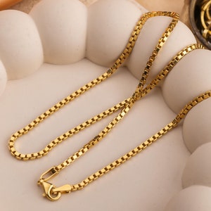 Puede incluir: Un collar de cadena de oro con un diseño de cadena de caja. La cadena está sobre una superficie blanca texturizada.