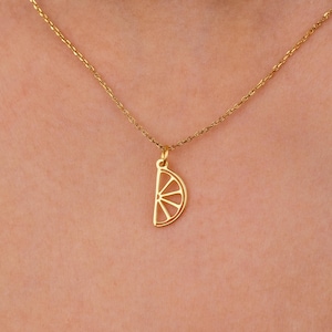 May include: A gold-colored necklace featuring a pendant shaped like a citrus fruit slice. The pendant has a detailed, open design, and hangs from a delicate chain. The necklace is worn against a skin tone background.