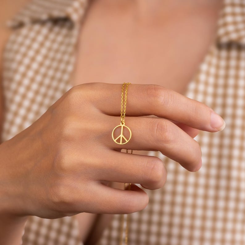 May include: A gold-colored necklace featuring a peace symbol pendant. The pendant is a simple, open design of the peace symbol. The necklace is displayed against a neutral background.