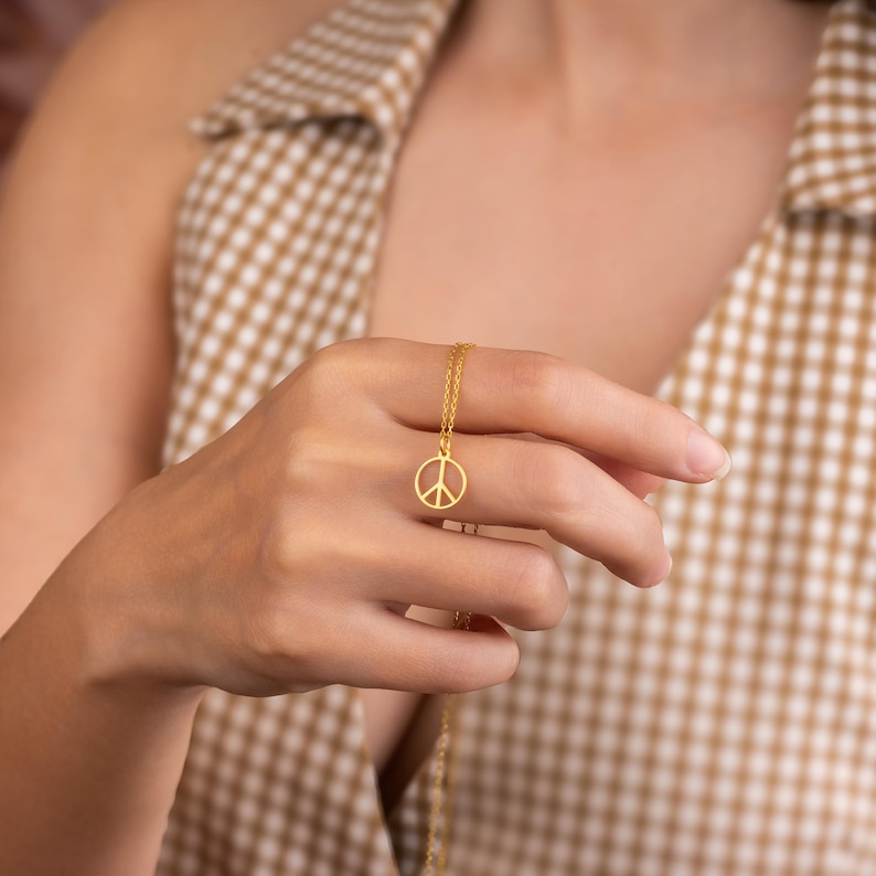 May include: A gold-colored necklace featuring a peace symbol pendant. The pendant is a simple outline of the peace sign, suspended from a delicate chain. The necklace is held up against a person's hand, with a checkered pattern in the background.