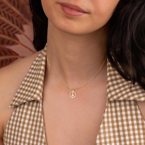 May include: A delicate gold-colored necklace featuring a peace symbol pendant. The pendant is a simple, open design. The necklace is worn against a backdrop of a brown and white checkered pattern.