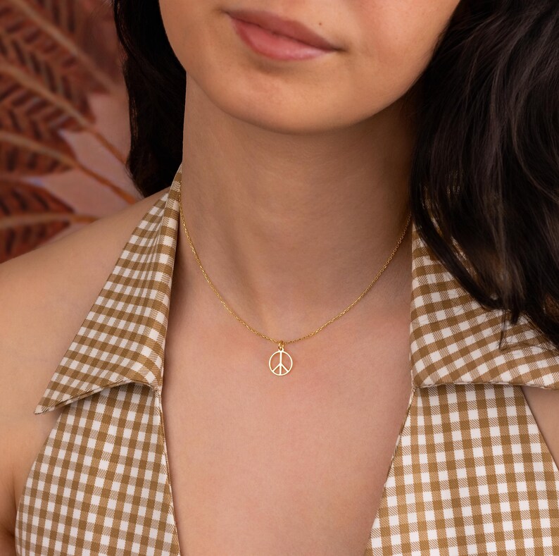 May include: A delicate gold-colored necklace featuring a peace symbol pendant. The pendant is a simple, open design. The necklace is worn against a backdrop of a brown and white checkered pattern.