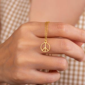 May include: A gold-colored necklace featuring a peace symbol pendant. The pendant is a circular outline with a vertical line and two diagonal lines inside. The necklace chain is delicate and also gold-colored.