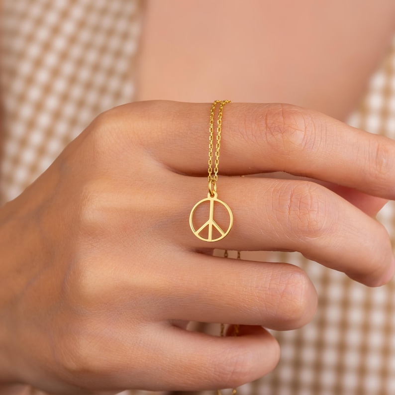 May include: A gold-colored necklace featuring a peace symbol pendant. The pendant is a circular outline with a vertical line and two diagonal lines inside. The necklace chain is delicate and also gold-colored.
