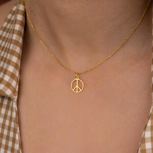 May include: A gold-colored necklace featuring a peace symbol pendant. The pendant is a simple, open design with a circle and three lines. The necklace is worn against a background of a person wearing a brown and white checkered shirt.