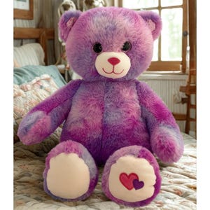 May include: A large, plush teddy bear in shades of purple and pink. The bear has a light pink muzzle, a red nose, and black eyes. The bear's paws have light pink pads, with one paw featuring two embroidered hearts. A cuddly toy.