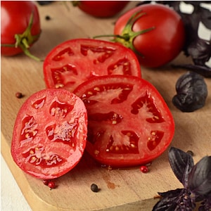 May include: Four slices of red ripe tomato on a wooden cutting board with black peppercorns and purple basil leaves.