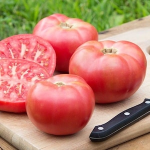 May include: Four large, ripe pink tomatoes on a wooden cutting board. Two tomatoes are sliced in half, revealing the red flesh and seeds. A black handled knife is in the foreground.