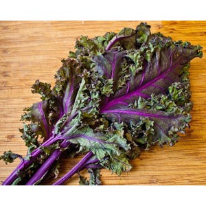 May include: A close-up of fresh red kale leaves with green edges and purple veins. The kale is on a wooden cutting board. The leaves have a ruffled texture.