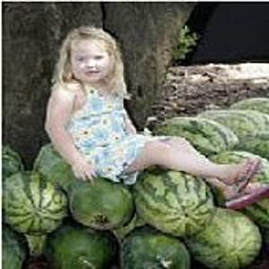 May include: A young girl sits on a pile of watermelons. The watermelons are green and have dark green stripes. The girl is wearing a light blue and white floral dress.
