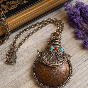 May include: A round, bronze-toned metal pendant shaped like a small flask, suspended from a matching chain. The pendant features intricate wirework, a decorative cap with inlaid turquoise and coral-colored stones, and etched script on the surface.